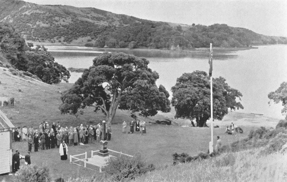 Black and white photograph of a group of people having a service in front of a marae on a grass stretch that runs down to the beach